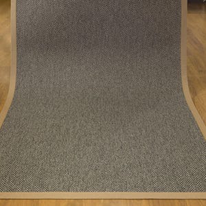May include: A rectangular area rug with a herringbone pattern in shades of gray and brown. The rug has a tan border and is displayed on a hardwood floor. The rug is made of a woven material.