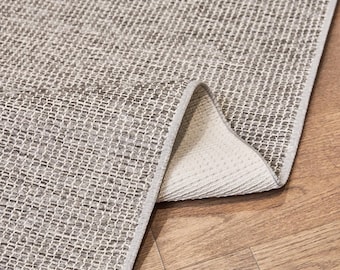 Puede incluir: Una alfombra de área de tonos neutros con una textura tejida. La superficie de la alfombra es una mezcla de hilos grises claros y oscuros, creando un patrón sutil. Una esquina de la alfombra está doblada hacia atrás, revelando una parte inferior blanca y un borde con flecos.
