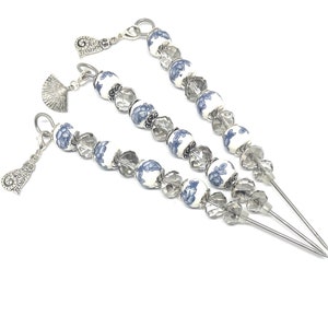 Puede incluir: Tres pinchos de plata y azul con cuentas y abalorios decorativos. Las cuentas son blancas con un patrón floral azul. Los abalorios son un abanico, un corazón y un remolino.