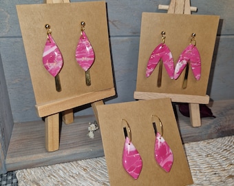 Könnte beinhalten: Präsentation handgefertigter Ohrringe in verschiedenen Stilen. Die Ohrringe werden auf kleinen Staffeleien präsentiert. Die Ohrringe haben ein marmoriertes Design in Rosa und Weiß mit goldfarbenen Akzenten. Die Ohrringe werden auf braunem Karton präsentiert.