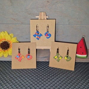Op de afbeelding: Presentatie van handgemaakte oorbellen op kleine kartonnen standaards. De oorbellen hebben kleurrijke, abstracte ontwerpen in blauw, rood, geel en groen, met goudkleurige hoepels. Een zonnebloem- en een watermeloenversiering zijn ook zichtbaar.