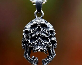 May include: A silver-colored pendant featuring a stacked skull design. The pendant is suspended from a silver chain. The skull has detailed features, including eye sockets, a nose cavity, and an open mouth. The background is blurred.