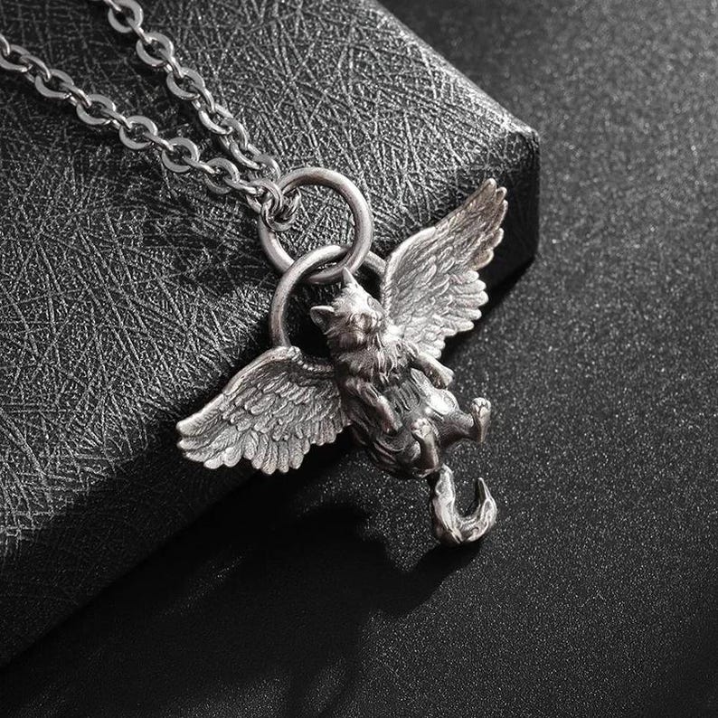 May include: A silver-colored pendant necklace featuring a winged cat design. The cat has detailed wings and is suspended from a chain. The pendant is intricately detailed and has a vintage aesthetic.