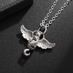 May include: A silver-colored pendant necklace featuring a winged creature design. The pendant has detailed wings and a curled tail, suspended from a delicate chain. The necklace is displayed against a dark background.