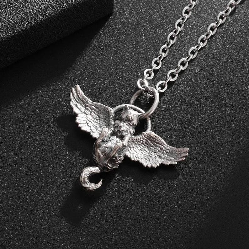 May include: A silver-colored pendant necklace featuring a winged creature design. The pendant has detailed wings and a curled tail, suspended from a delicate chain. The necklace is displayed against a dark background.