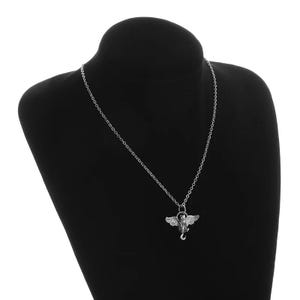 May include: Silver-toned necklace featuring a pendant with a winged creature design. The pendant is suspended from a delicate chain and displayed on a black mannequin. The necklace is a gothic or fantasy-themed jewelry item.