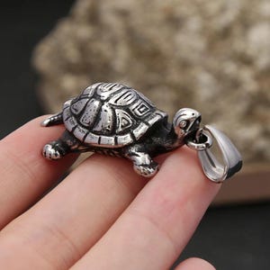 Könnte beinhalten: Ein silberfarbener Schildkrötenanhänger mit detailliertem Schalenmuster. Der Anhänger, mit einer Schlaufe für eine Kette, besteht aus glänzendem Metall. Kopf und Beine der Schildkröte sind sichtbar, was ihm ein dreidimensionales Aussehen verleiht.
