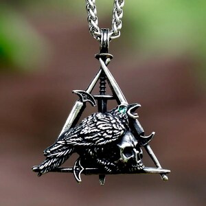 Puede incluir: Collar con colgante plateado que presenta un cuervo posado sobre una calavera dentro de un marco triangular. El cuervo tiene plumas detalladas y un ojo de gema verde. Dos espadas y una cadena completan el diseño. El collar cuelga de una cadena plateada.