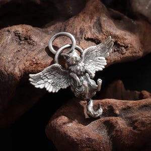 May include: A silver-colored pendant featuring a cat with feathered wings, suspended from a ring. The cat is depicted in a reclining pose, with its wings spread. The pendant is detailed and appears to be made of metal.