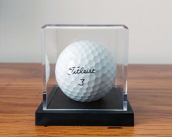 May include: A white golf ball with the text "Titleist 3" printed on it, displayed in a clear plastic cube with a black base.