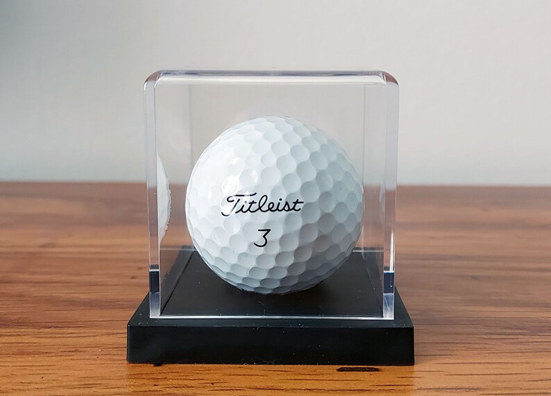 May include: A white golf ball with the text "Titleist 3" printed on it, displayed in a clear acrylic cube on a black base.