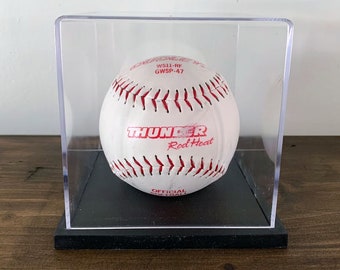 May include: A white softball with red stitching and the words "Thunder Red Heat" and "Official Softball" printed on it. The softball is displayed in a clear plastic display case with a black base.