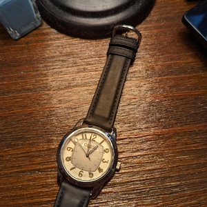 May include: A silver-toned wristwatch with a black leather strap. The watch face has a white background with black Roman numeral hour markers and black hands. The watch face also has a small date window at the 3 o'clock position. The watch is on a wooden surface.