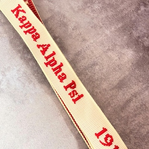 May include: Cream-colored lanyard with the text "Kappa Alpha Psi" in red, along with the number "19". The lanyard has a red trim along the edges. The text is clearly embroidered on the fabric.