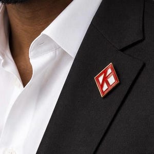 May include: A diamond-shaped lapel pin featuring a red and white geometric design with a gold-toned metal border, attached to the lapel of a black blazer worn over a crisp white collared shirt with visible buttons and a structured collar.