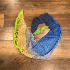 May include: A triangular knitted shawl in shades of green, blue, and brown. The shawl is folded in a spiral shape.