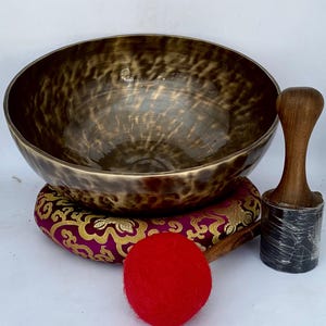 May include: A large, brown singing bowl with a wooden striker and a red felt mallet. The bowl is sitting on a purple and gold cushion.
