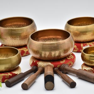 Pode incluir: Uma coleção de taças de canto de latão de vários tamanhos, apoiadas em almofadas estampadas em vermelho e dourado. Malhos de madeira são dispostos em frente às taças. As taças têm uma textura martelada e um tom dourado quente.