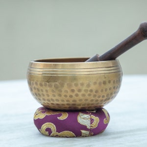May include: A gold singing bowl with a hammered texture sits on a purple cushion with gold accents. A wooden striker rests on the rim of the bowl.