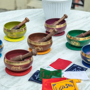 May include: Seven singing bowls with wooden strikers, each bowl is a different colour: blue, red, green, brown, purple, yellow, and a second blue bowl. The bowls are arranged on a white table with colourful fabric squares.