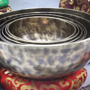 Puede incluir: Un juego de seis cuencos cantores de color bronce de varios tamaños, anidados unos dentro de otros. Los cuencos están sobre un cojín rojo con bordados dorados. Otros cojines y una maza son visibles en el fondo.