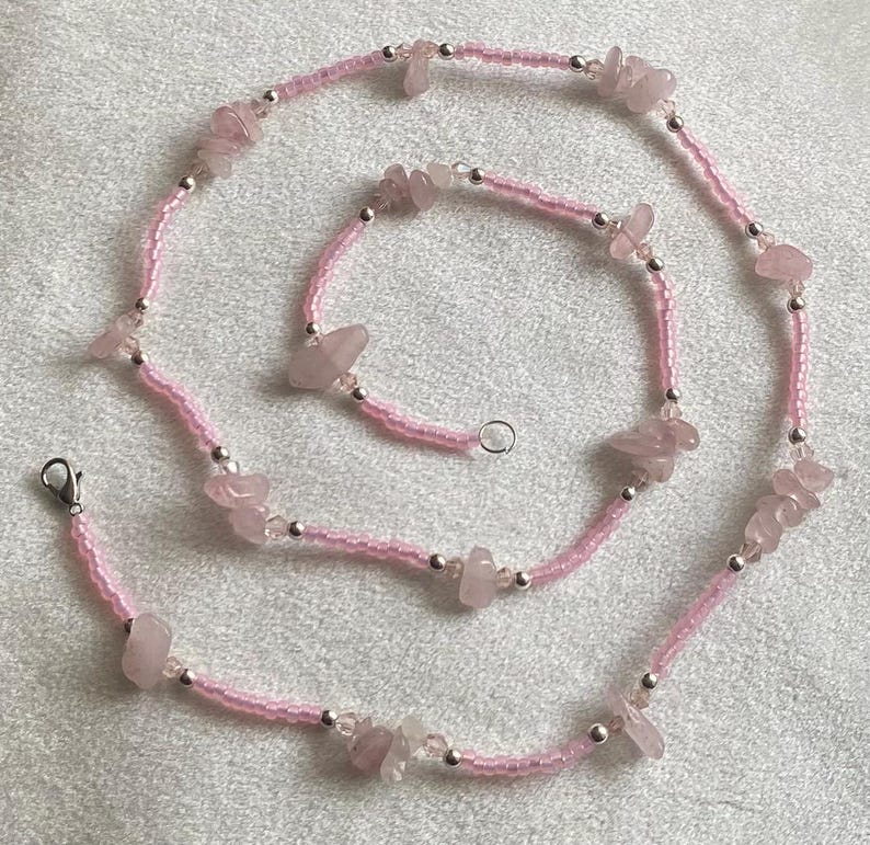 Puede incluir: Un collar de cuentas rosadas con piedras de cuarzo rosa. El collar est&aacute; hecho de peque&ntilde;as cuentas rosadas y tiene un cierre plateado.