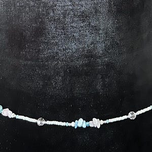 Op de afbeelding: Een delicate tailleband, met een witte kralenketting versierd met heldere, lichtblauwe en roze edelsteenaccenten. De kralen zijn aan elkaar geregen, waardoor een subtiel en elegant accessoire ontstaat. De tailleband wordt getoond op een zwart fluwelen oppervlak.