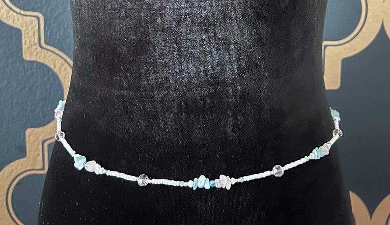 Op de afbeelding: Een delicate tailleband, met een witte kralenketting versierd met heldere, lichtblauwe en roze edelsteenaccenten. De kralen zijn aan elkaar geregen, waardoor een subtiel en elegant accessoire ontstaat. De tailleband wordt getoond op een zwart fluwelen oppervlak.