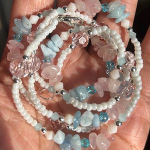 Op de afbeelding: Een meerstrengs ketting met witte zaadkralen, roze en blauwe edelstenen en heldere kristallen kralen. De ketting heeft een zilverkleurige sluiting en accenten. De edelstenen lijken rozenkwarts en aquamarijn te zijn.