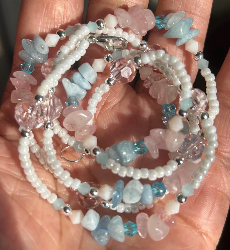 Op de afbeelding: Een meerstrengs ketting met witte zaadkralen, roze en blauwe edelstenen en heldere kristallen kralen. De ketting heeft een zilverkleurige sluiting en accenten. De edelstenen lijken rozenkwarts en aquamarijn te zijn.