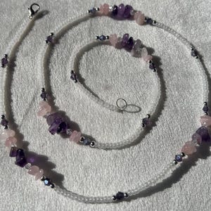 May include: A delicate necklace and bracelet set featuring a white beaded chain. The necklace and bracelet are adorned with amethyst and rose quartz gemstone chips, interspersed with small silver-toned beads and crystals. The jewelry is displayed on a white surface.