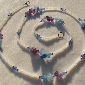 May include: A white beaded necklace with a silver clasp and various colored gemstone beads. The beads are shades of blue, purple, and light blue.
