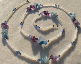 Op de afbeelding: Een witte kralenketting met een zilveren sluiting en verschillende gekleurde edelsteenkralen. De kralen zijn in tinten blauw, paars en lichtblauw.