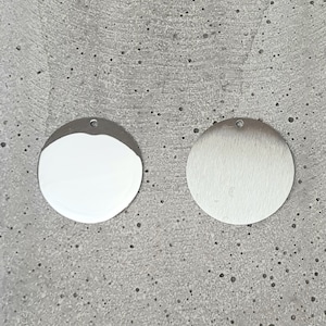 May include: Two round metal charms, one with a polished finish and the other with a brushed finish. Both charms have a small hole at the top for attaching to a necklace or bracelet.