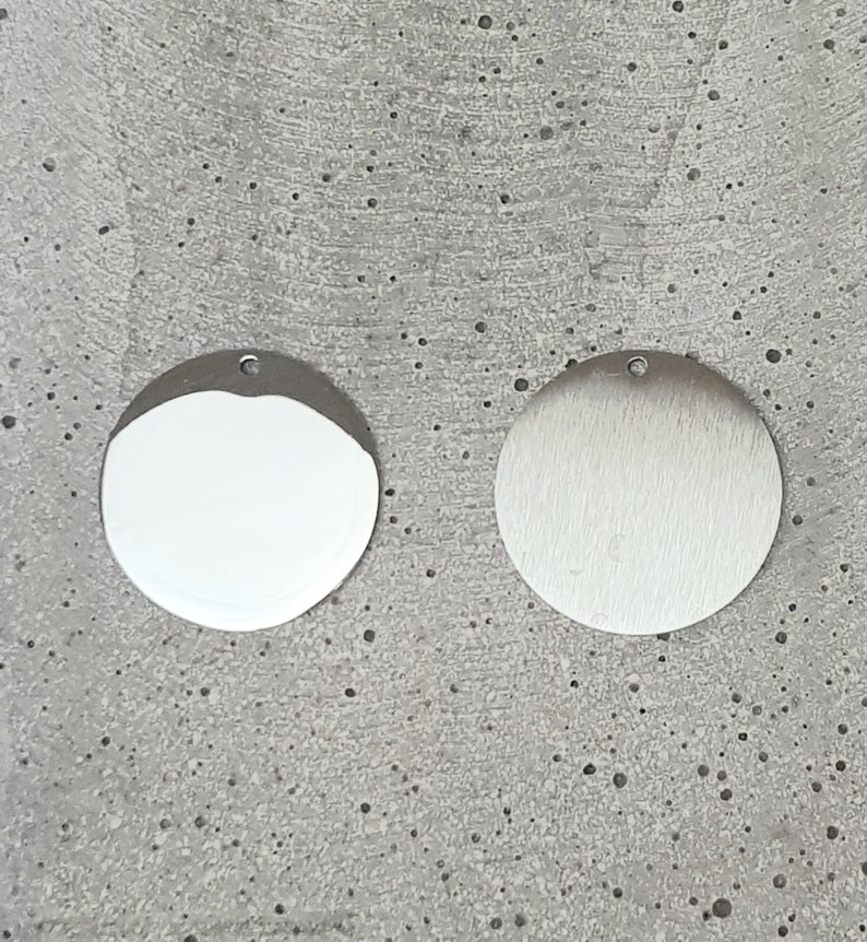 May include: Two round metal charms, one with a polished finish and the other with a brushed finish. Both charms have a small hole at the top for attaching to a necklace or bracelet.