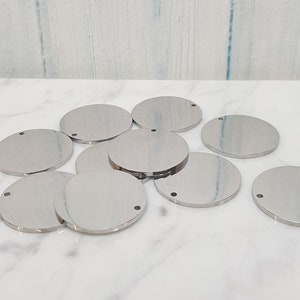 May include: A group of ten silver-colored metal circles with a small hole in the center. The circles are arranged in a pile on a white marble surface.