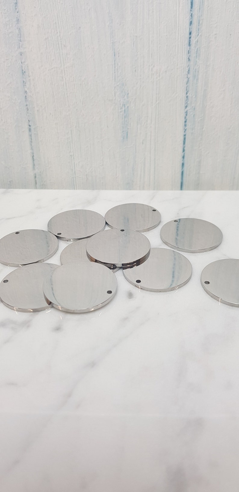 May include: A group of ten silver-colored metal circles with a small hole in the center. The circles are arranged in a pile on a white marble surface.