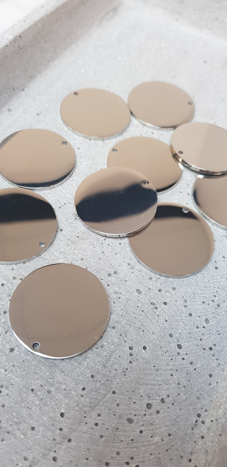 May include: A group of silver-colored metal circles with a small hole in the center. The circles are arranged in a random pattern on a gray textured surface.