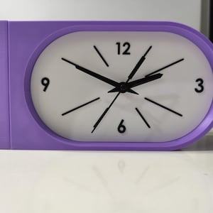 Puede incluir: Un reloj de escritorio horizontal de forma ovalada con un marco de color morado vibrante y una esfera blanca. El reloj presenta números negros en las posiciones de las 3, 6, 9 y 12, con manecillas negras para horas, minutos y segundos. El diseño incluye una sección rectangular en el lado izquierdo.