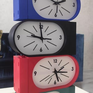 Peut inclure: Une pile verticale de quatre horloges analogiques rectangulaires aux bords arrondis. Chaque horloge présente un cadran blanc avec des marqueurs d'heure noirs, des aiguilles noires et les chiffres 3, 6, 9 et 12. Les cadres sont bleu, noir, rouge et bleu sarcelle.