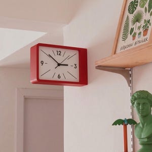 May include: A rectangular wall clock with a vibrant red frame and a white face. The clock features black hour markers, black hands, and black numerals at the 3, 6, 9, and 12 o'clock positions. The design is clean and functional, mounted directly on a wall.