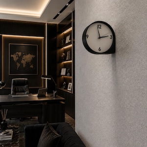 May include: A round wall clock with a black frame and a white face featuring black numerals at the 12, 3, 6, and 9 positions. The clock has two black hands and is mounted on a textured light gray wall in a room with dark office furniture.
