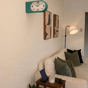 May include: A double-sided wall clock with a teal casing and a white face featuring black numerals at 3, 6, 9, and 12 o'clock. The clock has black hour and minute hands and is mounted on a wall above a sofa, side table, and floor lamp.