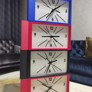May include: A vertical stack of four rectangular clocks with white faces and black hands. Each clock features black numerals at the 3, 6, 9, and 12 positions, with black radial lines. The frames are coloured blue, red, black, and pink from top to bottom.