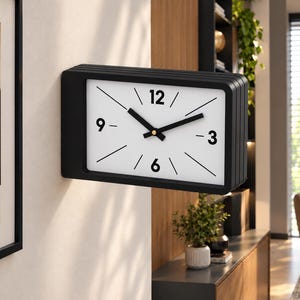 Puede incluir: Un reloj de pared rectangular con marco negro mate y esfera blanca. El reloj presenta números negros en negrita en las posiciones de las 12, 3, 6 y 9, con líneas negras finas que marcan las horas restantes y manecillas negras de hora y minuto.