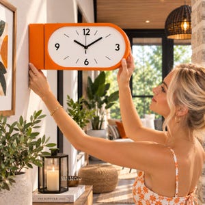 Peut inclure: Une horloge murale horizontale de forme ovale avec un cadre orange vif et un cadran blanc. L'horloge présente des chiffres noirs aux positions 3, 6, 9 et 12 heures, avec des traits noirs simples pour les autres heures et des aiguilles noires.