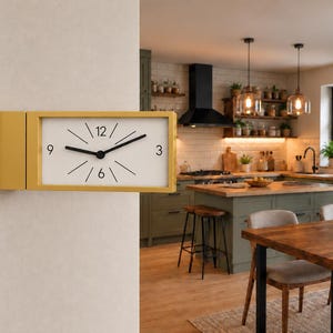 May include: A rectangular wall clock with a mustard yellow frame and a white face. The clock features black hour and minute hands, black numerals at the 3, 6, 9, and 12 positions, and thin black lines representing the remaining hour markers.