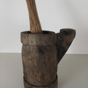 May include: A wooden mortar and pestle set. The mortar is brown and has a handle on the side. The pestle is also brown and has a rounded end.