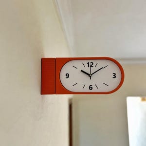 May include: A wall-mounted clock with an orange, pill-shaped frame and a white face. The clock features black hour and minute hands, with black numerals at the 12, 3, 6, and 9 o'clock positions, and simple black tick marks for the remaining hours.