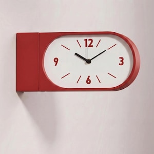 May include: A red wall clock with a white oval clock face. The clock has black hands and red numerals at the 12, 3, 6, and 9 o'clock positions. The clock is mounted on a white wall.